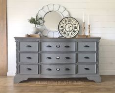 a gray dresser with drawers and a clock on it's top, in front of a white wall