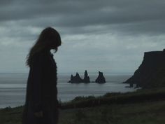 a woman standing on top of a lush green hillside next to the ocean under a cloudy sky