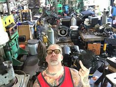 a man with tattoos and glasses sitting in a room full of machines, tools and other items