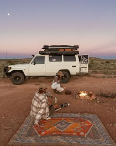 two men sitting on the ground next to a campfire with an suv in the background