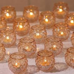 many small glass candles are lined up on a white tablecloth with the lights turned on