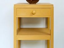 a small yellow table with a bowl on it's top, against a white wall