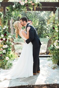 a bride and groom kissing in front of an outdoor ceremony arch with flowers on it