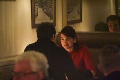 a woman sitting at a table next to a man in a red shirt and black jacket