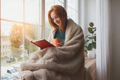 a woman sitting on a window sill reading a book and holding a coffee cup