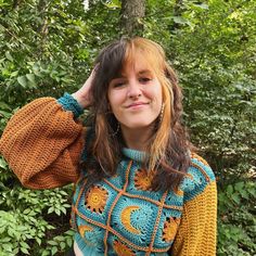 a woman with long hair wearing a crocheted sweater in front of some trees