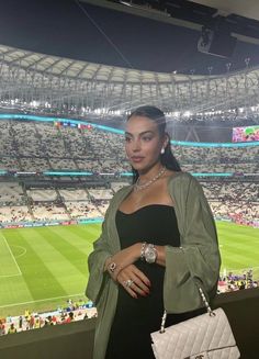 a woman standing in front of a stadium holding a white purse and looking at the camera