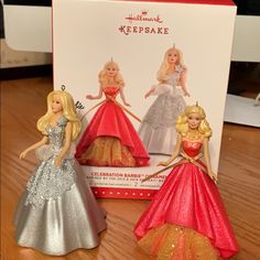 three barbie dolls are sitting in front of a box on the table, and one is wearing a dress with sequins