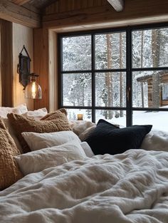 a bed with white sheets and pillows in front of a window that looks out onto the snow covered forest