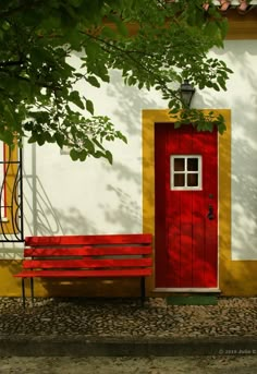 a red door and bench in front of a white building with yellow trim on it