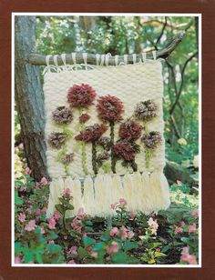 a crocheted wall hanging with flowers on it in the grass next to a tree