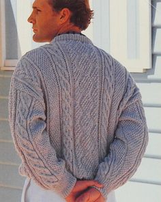 a man wearing a gray cabled sweater and white pants standing in front of a house