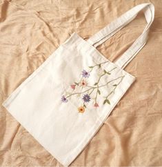 a white bag with flowers embroidered on it sitting on a bed sheet, next to a pillow