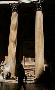 people are walking around in an old building with columns and paintings on the wall behind them