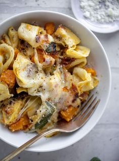 a white bowl filled with pasta covered in cheese and sauce next to a fork on top of a table