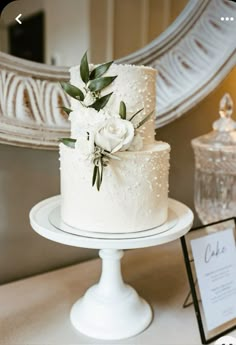 a wedding cake with white flowers and greenery sits on a table in front of a mirror