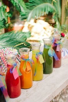 four jars filled with different colored drinks on top of a wooden table next to palm trees