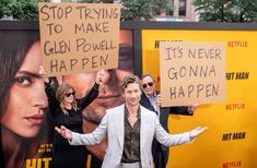 a man standing in front of a movie poster holding up two signs that say stop trying to make glenn powell happen