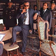 a man in a suit and tie sitting on a chair with other men around him