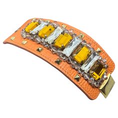 This bracelet by Gedebé showcases a bold, opulent design. It features a curved, textured orange leather band adorned with a striking array of rectangular gemstones. The central stones are large, faceted yellow gems, framed by clear crystals that create a dazzling effect. The bracelet is further accented by small, gold-tone pyramid studs along the edges, adding a touch of edginess to the overall piece. The vibrant colors and mix of textures make this bracelet a statement accessory, combining luxu Yellow Gems, Orange Texture, Gems Bracelet, Orange Leather, Clear Crystals, Brown Skin, Leather Band, Clear Crystal, Contemporary Style