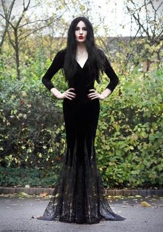 a woman with long hair wearing a black dress standing in front of bushes and trees