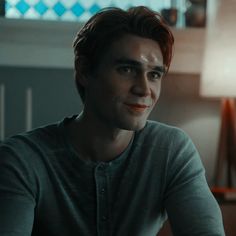 a young man sitting in front of a lamp looking at the camera while wearing a grey shirt