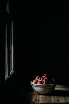 a bowl full of strawberries sitting on a table next to a window in a dark room