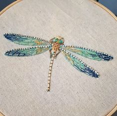 a blue dragonfly sitting on top of a white cloth in a wooden hoop with beading around it