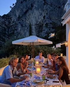 a group of people sitting around a table with food and drinks in front of a mountain