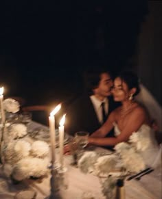 a bride and groom sitting at a table with candles in front of their faces, surrounded by white flowers