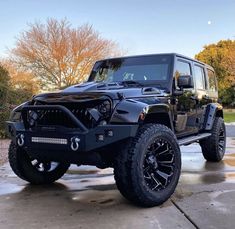 a black jeep parked on top of a parking lot in front of some trees and bushes