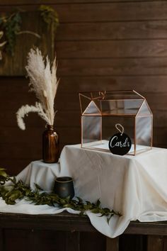 a table topped with a small glass box next to a vase filled with flowers and greenery