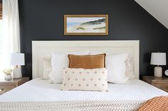 a white bed topped with pillows next to two lamps and a painting on the wall