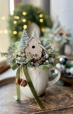 a white mug with a birdhouse in it on top of a wooden table next to a christmas tree