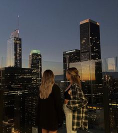 two women standing on top of a tall building looking at the city skyline with skyscrapers in the background