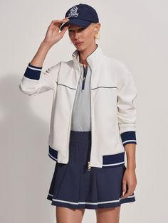 a woman in a white jacket and blue skirt is holding her hat to her head