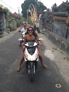 two people riding on the back of a motor scooter down a narrow street