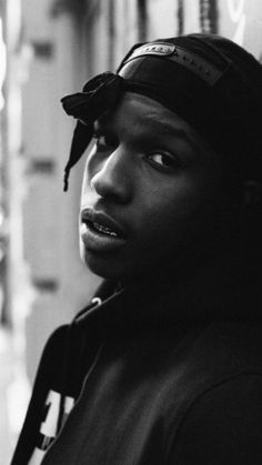 a black and white photo of a young man leaning against a wall wearing a hat