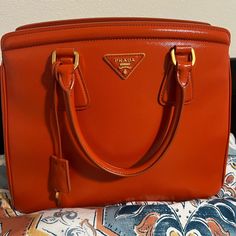 Very Good Condition Prada Handbag. Please Check Photos For Condition. Good Size Handbag For All Your Essentials. Only Callouts Are Black Ink Stain In The Interior Pocket And Some Minor Surface Cracks In One Corner Opening As Shown In Photos. Designer Orange Office Bags, Luxury Orange Office Bag, Luxury Orange Handheld Bag, Luxury Handheld Orange Bag, Designer Orange Satchel, Designer Orange Satchel For Shopping, Classic Orange Bag With Gold-tone Hardware, Designer Orange Formal Bags, Designer Orange Bags For Formal Occasions