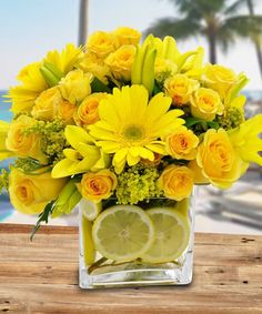 a vase filled with yellow flowers and lemons on top of a wooden table next to the ocean