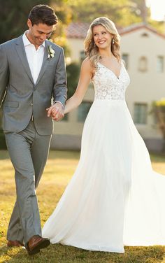 a bride and groom holding hands while walking through the grass in front of a house