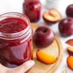 a person holding a jar of plum jam with sliced peaches around it on a cutting board