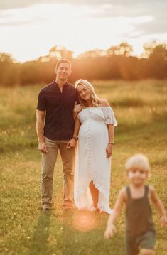 a pregnant woman standing next to a man in a field with a toddler walking behind her