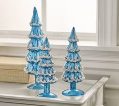 two blue glass christmas trees sitting on top of a white table next to a window