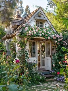 a small white house with pink flowers growing on the roof and door, surrounded by greenery