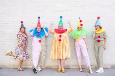 four women dressed in clown costumes posing against a brick wall with their hands up to the ceiling