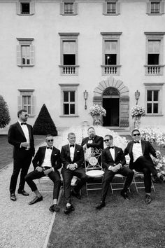 black and white photograph of men in tuxedos sitting on a bench outside a large building