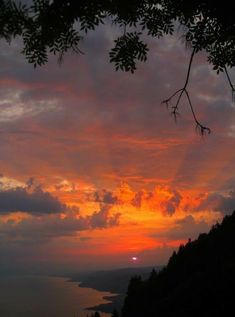 the sun is setting over some trees and water in the distance, with clouds above it