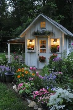 a small white shed with flowers in the front yard and lights on it's windows