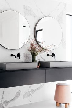 two round mirrors on the wall in a bathroom with marble counter tops and black sinks